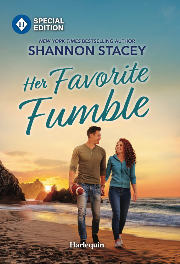 Cover of Her Favorite Fumble. A couple smiling at each other while holding hands and walking on a rugged, rocky beach with a sunset behind them. He has short dark hair and is wearing an olive Henley shirt and worn jeans. He’s holding a football in his free hand. She has long, curly red hair and is wearing a blue hoodie and jeans.